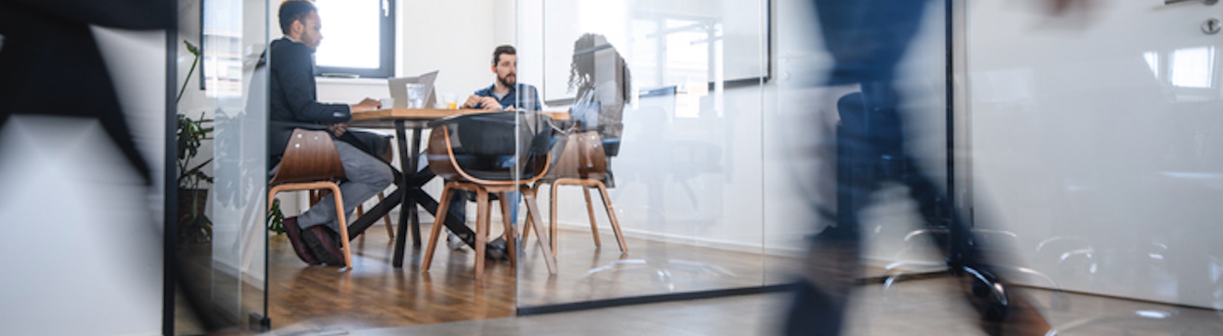 Three professionals in a modern glass-walled conference room engaged in a meeting, with a blurred figure walking by in the foreground, symbolizing a dynamic office environment.